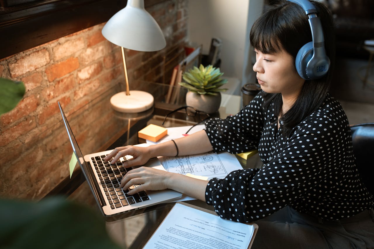 about-01 Focused woman wearing headphones, working on a laptop in a cozy home office setup with natural light.